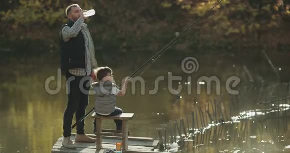 放松时间爸爸和他的爸爸在阳光明媚的日子在湖边钓鱼教儿子钓鱼爸爸喝一瓶水视频的预览图