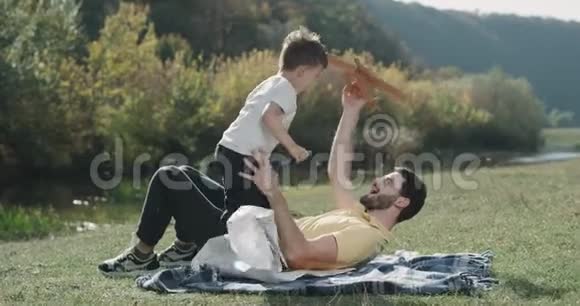 有魅力的三岁男孩在大自然中和爸爸玩飞机他们躺在草地上视频的预览图