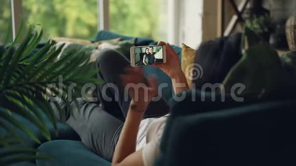 社交女孩用智能手机在舒适的房子里躺在沙发上打视频电话的后景人们在交谈视频的预览图