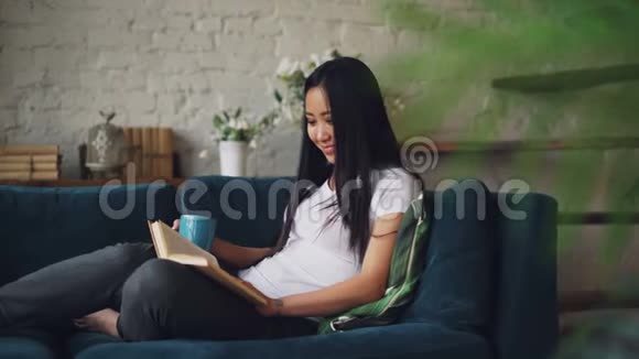 美丽的亚洲女人正在看书在舒适的阁楼式公寓里坐在沙发上享受着有趣的故事和欢笑视频的预览图