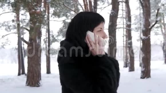 穿着毛皮外套的布鲁内特女士在冬天站在森林或公园里打电话世界上视频的预览图