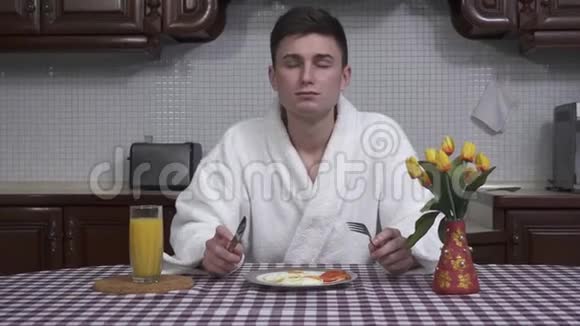 漂亮的年轻人穿着白色浴袍坐在厨房的桌子旁享受着早餐的香气煎锅视频的预览图