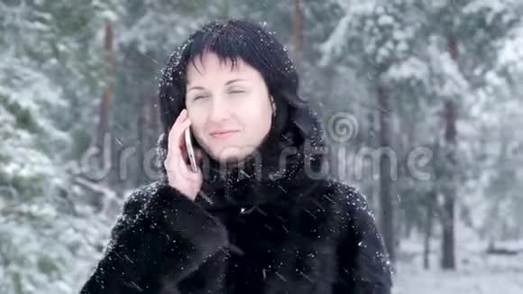 穿着黑色外套的布鲁内特女孩在冬天的公园或森林里打电话微笑着视频的预览图