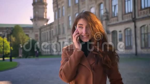 可爱的白种人黑发女孩站在反黑建筑附近用华丽的微笑和快乐的表情在电话中交谈视频的预览图