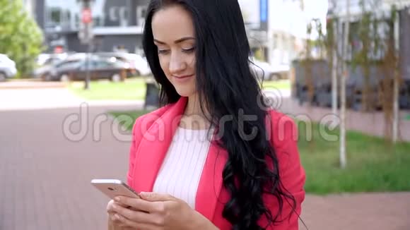 穿红色夹克的苗条女孩在夏天的公园里使用智能手机视频的预览图