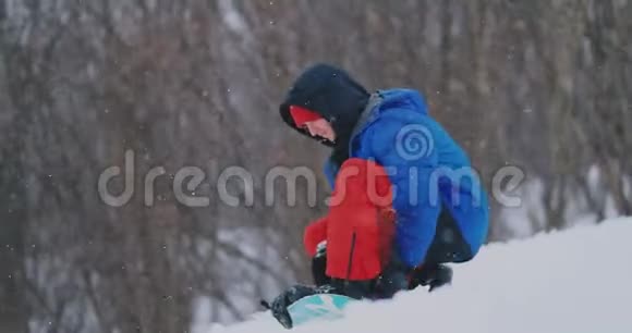 一个穿着红裤子的人坐在雪地上在滑雪坡上系着滑雪板鞋和一件蓝色夹克视频的预览图
