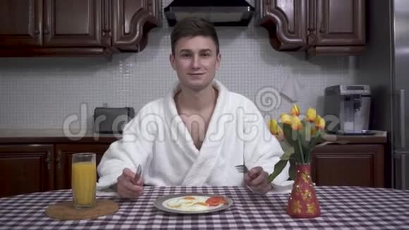 英俊的年轻人穿着白色浴袍坐在厨房的桌子旁享受着早餐的香气煎锅视频的预览图