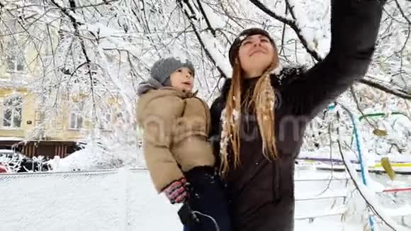 年轻母亲抱着她蹒跚学步的儿子摇动覆盖着雪的树枝的4k视频家庭娱乐视频的预览图
