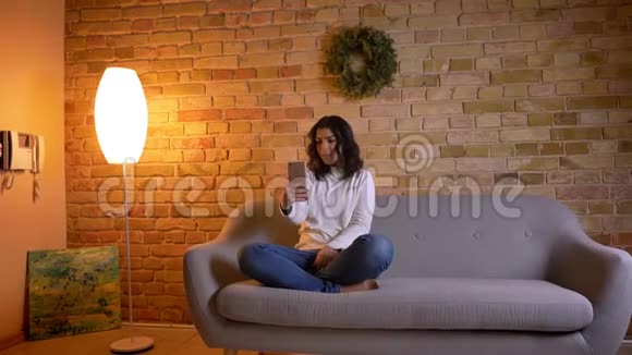 白种人黑发家庭主妇坐在沙发上在智能手机上愉快地在家里用视频聊天的肖像视频的预览图