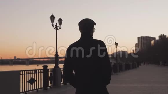 日落时分黄昏时分一个穿着外套的年轻人沿着河边的人行道散步从后面看视频的预览图