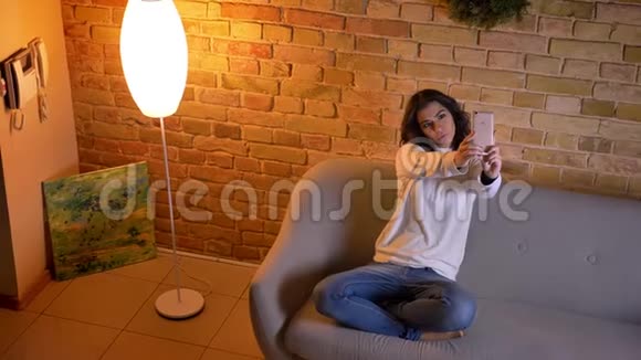 漂亮的黑发女商人坐在沙发上在家里愉快地在智能手机上制作自拍照片视频的预览图