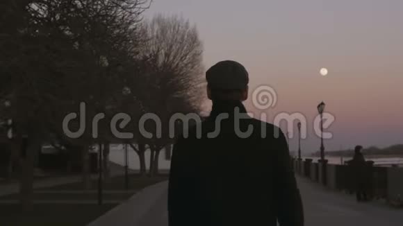 日落时分黄昏时分一个穿着外套的年轻人沿着河边的人行道散步从后面看视频的预览图