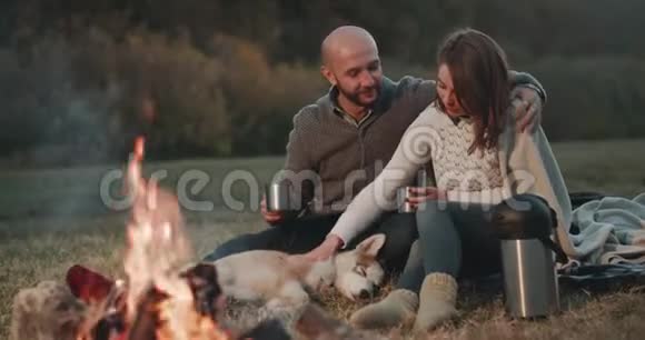 在大自然中美丽的一对夫妇和他们的哈士奇狗在篝火旁玩得很开心一起拿着一个铁杯和一个视频的预览图