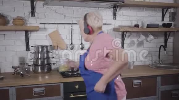 滑稽的白种人老妇人戴着耳机在厨房唱歌跳舞成熟开朗的退休者享受生活后视频的预览图