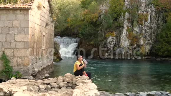 女人坐在瀑布前的岩石上喝水视频的预览图