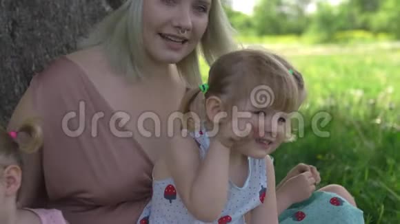 年轻的金发嬉皮妈妈和她的小女孩在公园的树下坐着笑视频的预览图