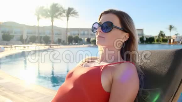 华丽美丽的女孩穿着性感的红色比基尼和太阳镜躺在酒店泳池边的躺椅上的肖像妇女视频的预览图