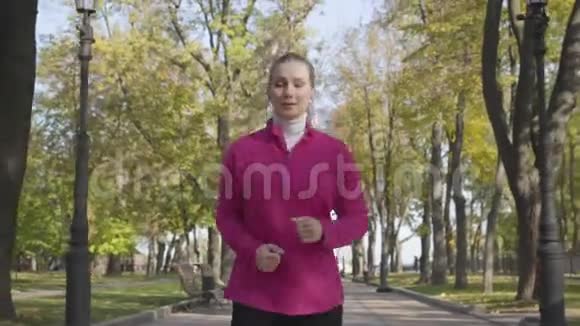 穿着运动服和耳机在秋天公园跑步的年轻白种人女人早上女子跑步训练视频的预览图