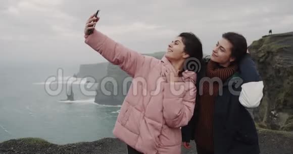 使用智能手机两位美丽的女士在悬崖顶的屋顶上拍照留念令人叹为观止视频的预览图
