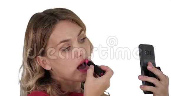 漂亮时尚的年轻女人在嘴唇上涂红色口红看着白色背景上的手机屏幕视频的预览图