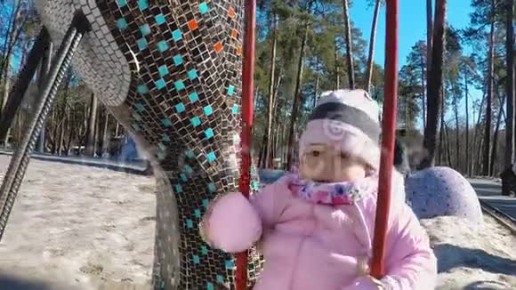 一个穿着粉红色夹克和条纹帽子的小女孩正骑着秋千在一个公园里早春有一棵高大的松树视频的预览图