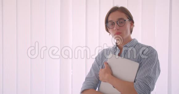 一位年轻英俊的白种人商人戴着一副眼镜手里拿着一台笔记本电脑站在办公室的特写肖像视频的预览图