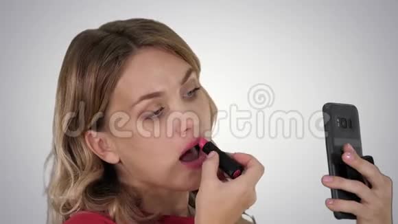 漂亮时尚的年轻女人在嘴唇上涂红色口红在渐变背景上看手机屏幕视频的预览图