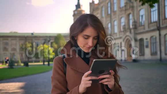 可爱的白种人黑发女孩站着放松双手拿着平板电脑看着它使用她的设备视频的预览图