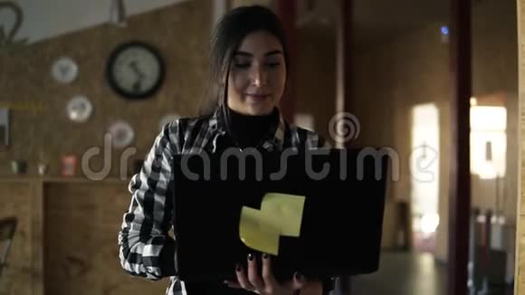 穿着格子衬衫的年轻黑发女郎正在现代工作空间工作带着笔记本电脑在房间里行走微笑视频的预览图