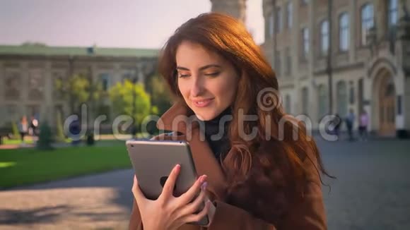 快乐的褐发白种人女孩正微笑着在平板电脑上的镜头下展示绿色的屏幕在户外阳光下视频的预览图