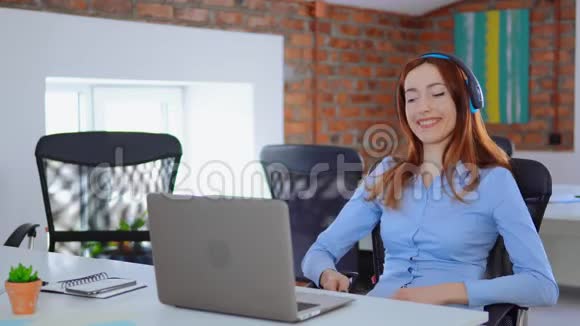 快乐的年轻女子边唱歌边用耳机听大声的音乐坐在办公桌旁用笔记本电脑视频的预览图