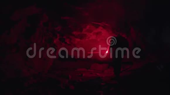 年轻人手里拿着一个燃烧的红色信号耀斑来到黑暗的地下洞穴的窄洞里股票视频的预览图