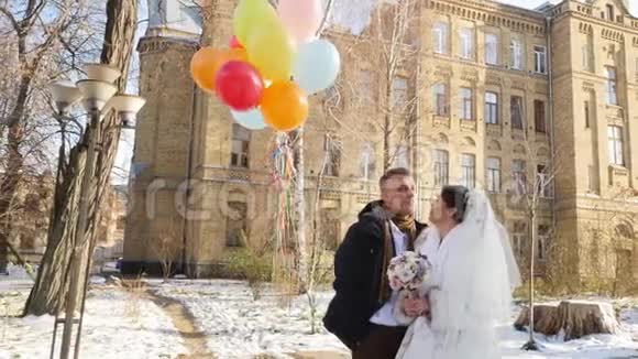 冬天的婚礼快乐欢笑的新婚夫妇带着明亮的多色的气球晴朗的雪天古老的建筑视频的预览图