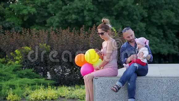 年轻的爸爸和可爱的小女儿坐在水泥的窗台上妈妈坐在旁边正在画城公园里打电话视频的预览图