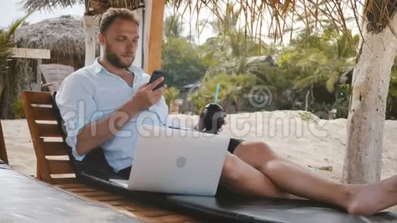 成功的快乐轻松商人与智能手机笔记本电脑和水果饮料坐在海滩躺椅度假视频的预览图