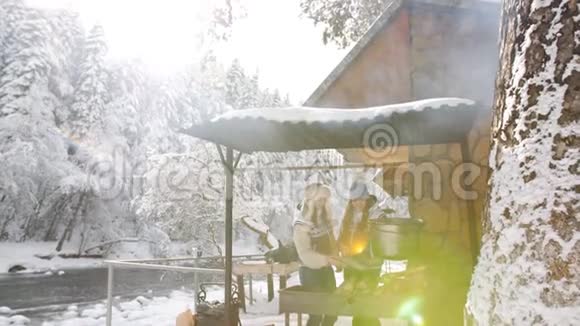 在一个明亮的冬天两个女人在靠近森林木制Chalet的炉子上准备食物视频的预览图