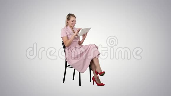 有趣的漂亮年轻女人穿着粉红色的连衣裙坐在椅子上在渐变背景上使用平板电脑视频的预览图