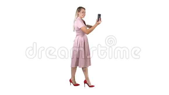 穿着粉色衣服的年轻女子在白色背景下行走时使用智能手机录制视频博客视频的预览图