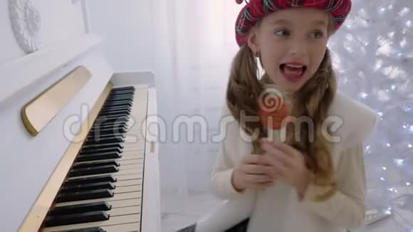 戴着红帽子的小女孩舔着白色钢琴和圣诞树附近的棒棒糖视频的预览图