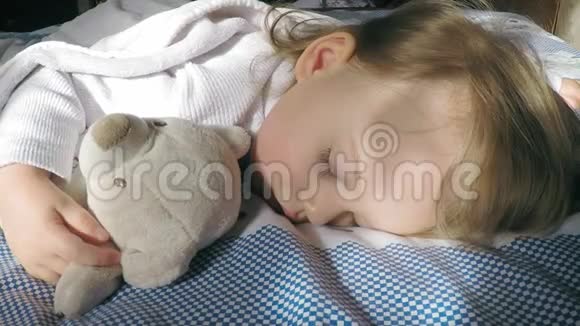 美丽的小女孩金发睡在床上被阳光照亮倒着一只泰迪熊视频的预览图