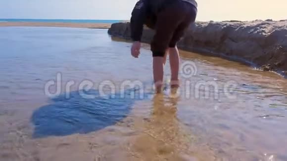 小男孩光着脚在流入大海的河流中的浅水中行走视频的预览图