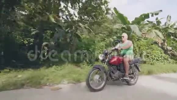 老人骑摩托车在热带村庄的道路上成熟男子驾驶摩托车而莫托旅行视频的预览图