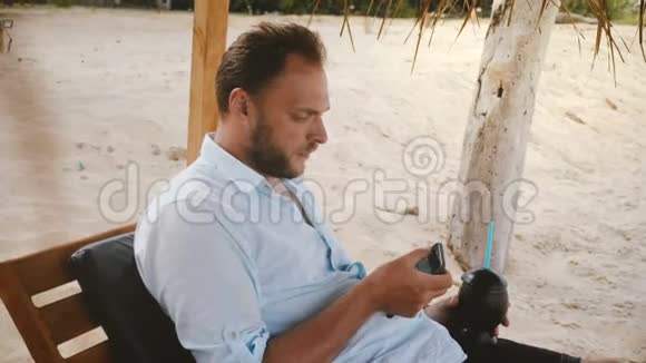 快乐自信的男性游客带着智能手表和水果鸡尾酒坐在沙滩椅上使用智能手机社交网络视频的预览图