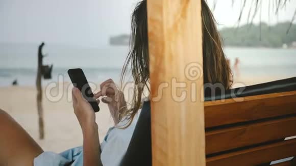 年轻女性使用智能手机购物应用程序在休闲海滩椅子上放松拥有令人惊叹的海滨风景视频的预览图