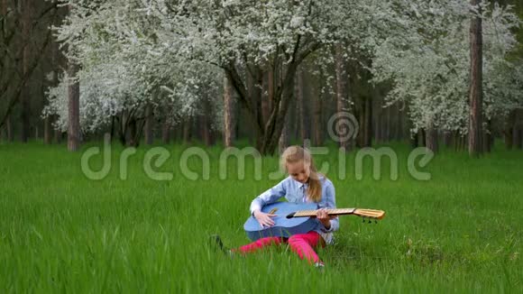 小女孩吉他手独奏坐在一个美妙的地方在一棵盛开的树绿草的背景上调你的吉他我视频的预览图