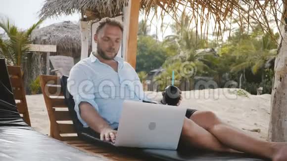 成功的中年男性自由职业者带着水果饮料在夏日海滩的躺椅上放松在笔记本电脑上打字视频的预览图