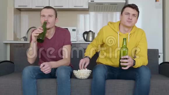 两个男人坐在沙发上看电视上的足球赛喝啤酒吃爆米花视频的预览图