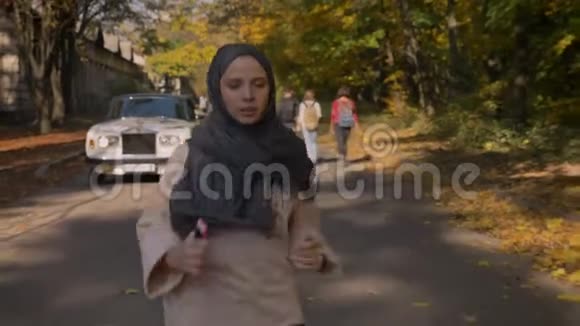 在秋色森林里年轻的穆斯林女孩戴着头巾带着手机在后台运行视频的预览图