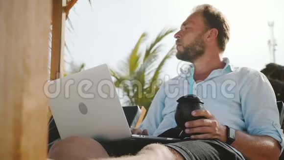 低角度拍摄成功快乐舒适自由职业者手提电脑和饮料休息海滩休闲椅视频的预览图