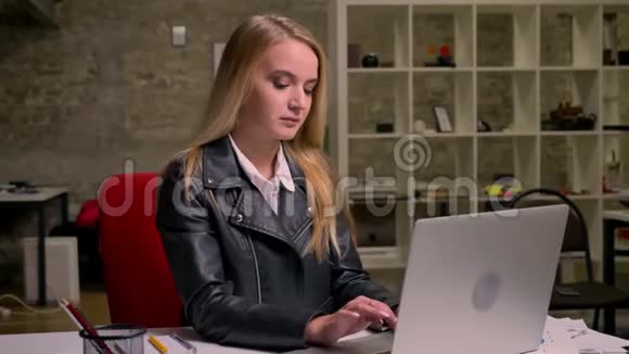 一位美丽的金发白种人女士正在用笔记本电脑打字然后看着相机在灯光下微笑视频的预览图